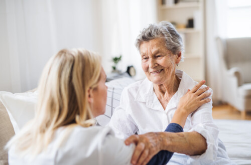 A health visitor talking to a sick senior woman sitting on bed at home.