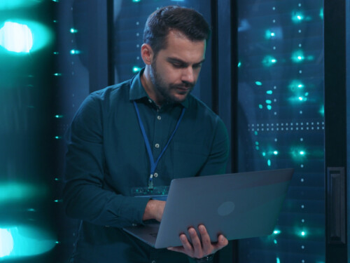 Caucasian IT professional admin using laptop computer doing data transfer operation with rack server cabinets in digital room of data center. Cyber security.