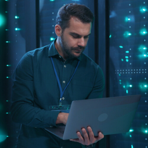 Caucasian IT professional admin using laptop computer doing data transfer operation with rack server cabinets in digital room of data center. Cyber security.