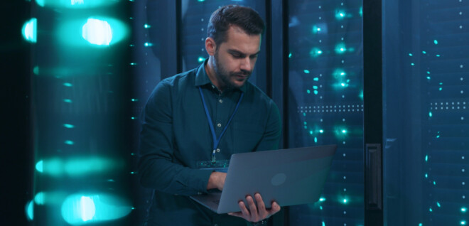 Caucasian IT professional admin using laptop computer doing data transfer operation with rack server cabinets in digital room of data center. Cyber security.