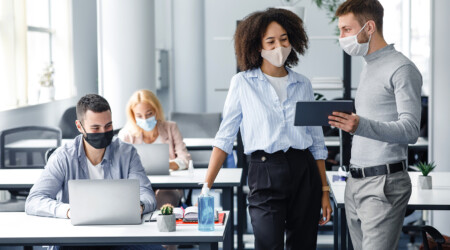 New project and plans for work. Boss in protective mask with tablet gives instructions to workers in masks, african american woman looking at gadget