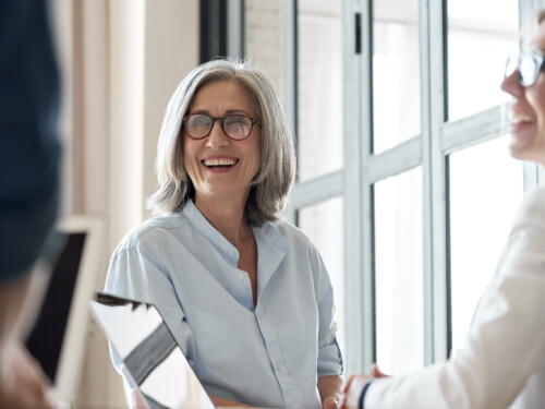 Happy old female mentor talking to young interns at group office meeting.