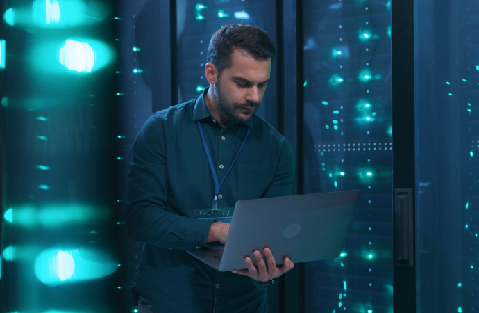 Caucasian IT professional admin using laptop computer doing data transfer operation with rack server cabinets in digital room of data center. Cyber security.