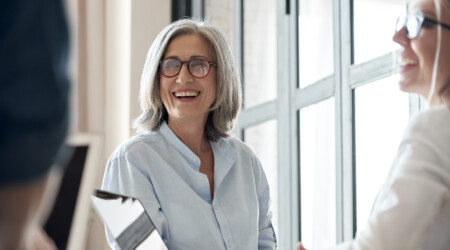 Happy old female mentor talking to young interns at group office meeting.