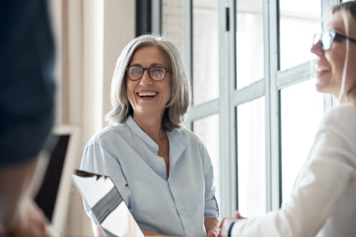 Happy old female mentor talking to young interns at group office meeting.