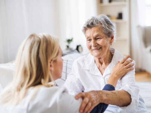 A health visitor talking to a sick senior woman sitting on bed at home.