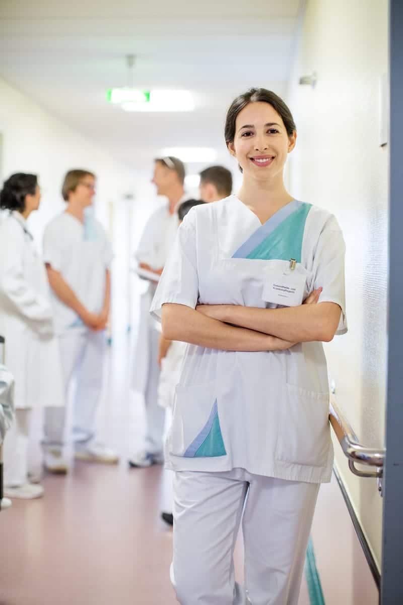 Infirmière souriante les bras croisés, avec une équipe médicale en arrière-plan dans le couloir.
