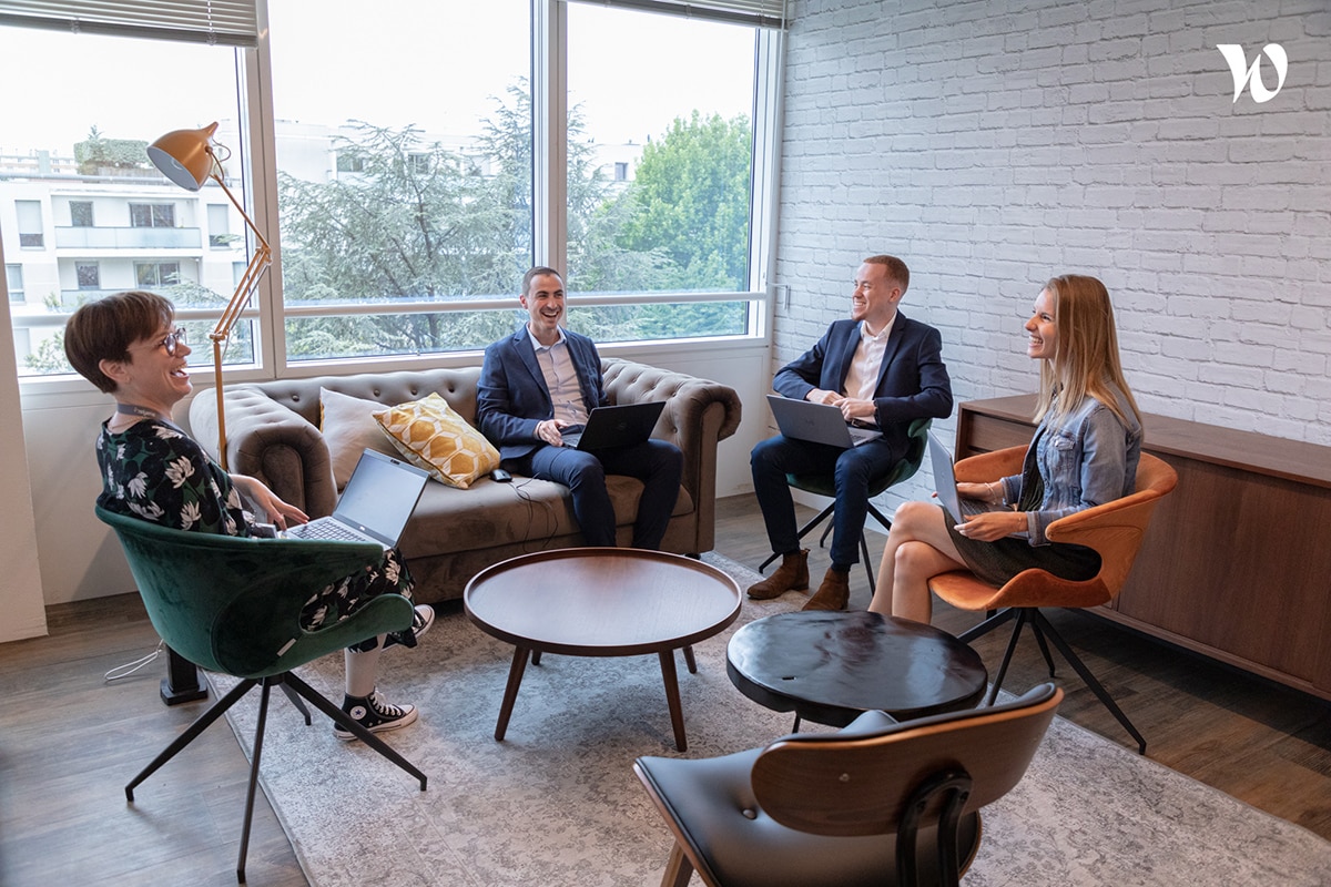 Quatre collaborateurs en réunion informelle et souriante dans un salon d'entreprise confortable.