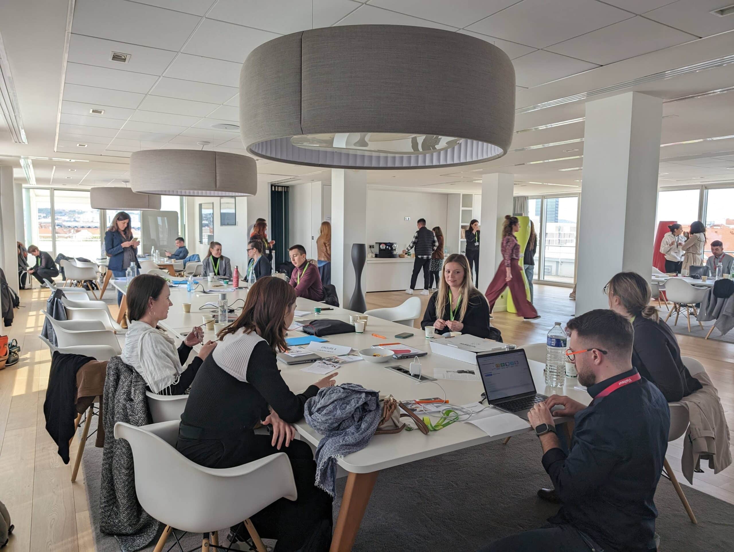 Grande salle de travail avec plusieurs groupes de collègues réunis autour de tables